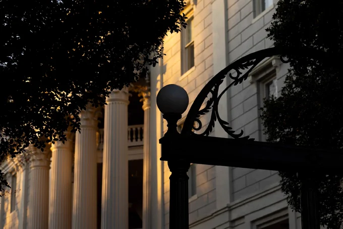 The image shows the University of Georgia Arch silhouetted in the foreground, with the sunlit columns of a classical building in the background, partially hidden by trees.