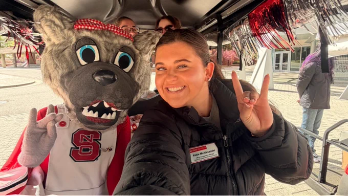 Person posing with Ms Wuf mascot wearing an NC State jersey inside a decorated golf cart, both making Wolfie hand signs.