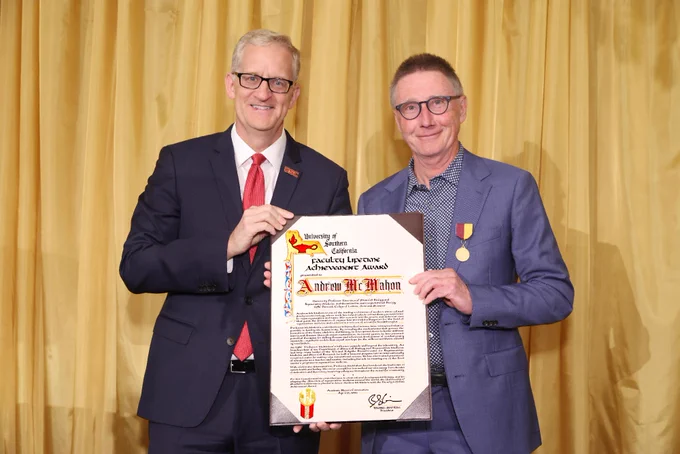 Two individuals stand side by side, smiling, with one holding a framed certificate titled "University of Southern California" and mentioning "Andrew McMahon." Both are dressed in formal attire, and one person is wearing medals on their jacket. A golden curtain serves as the backdrop.