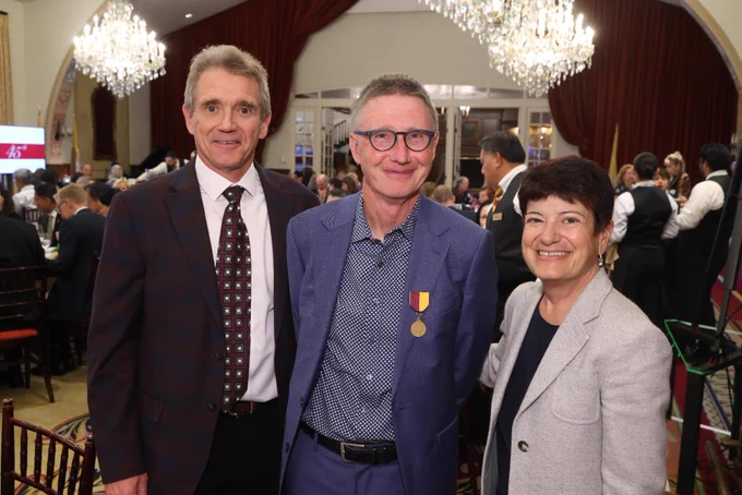 Three individuals posing for a photo at a formal sit down dinner banquet hall. The individual in the middle is wearing a medal on their jacket. 