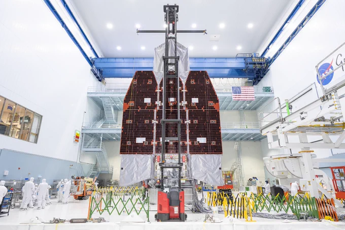 The Nancy Grace Roman Space Telescope with three large black solar panels deployed. The massive spacecraft is in a large open room with a viewing window on the wall on the left. There are extendable gates of different colors all around the telescope. A cherry-picker machine is fully extended in front of the telescope. Credit: NASA