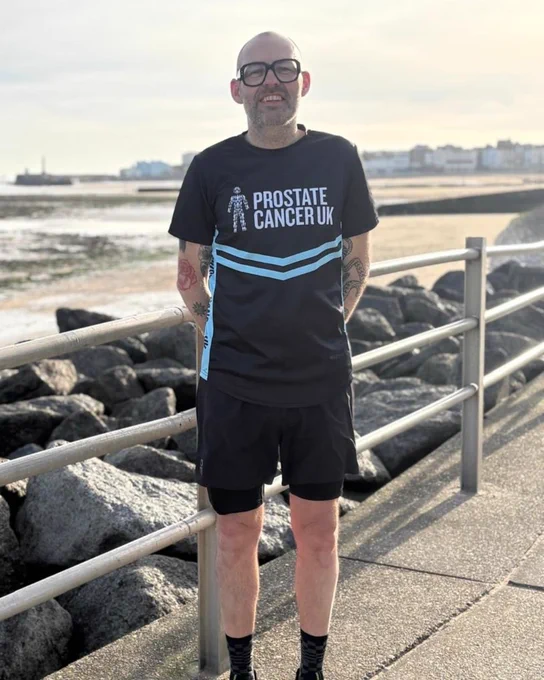 Chris standing on a boardwalk near a rocky shoreline, wearing a Prostate Cancer UK running t-shirt and shorts. There's a railing behind and buildings are visible in the distance under a partly cloudy sky.
