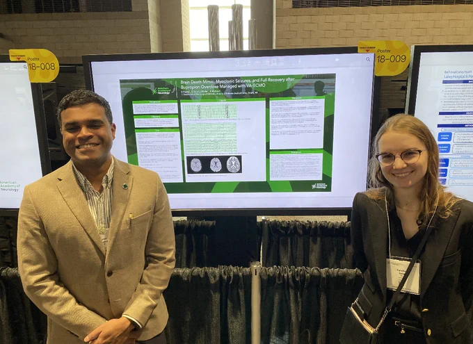 Drs. Sane and Mathew stand on either side of a large digital research poster. They are smiling to the camera.