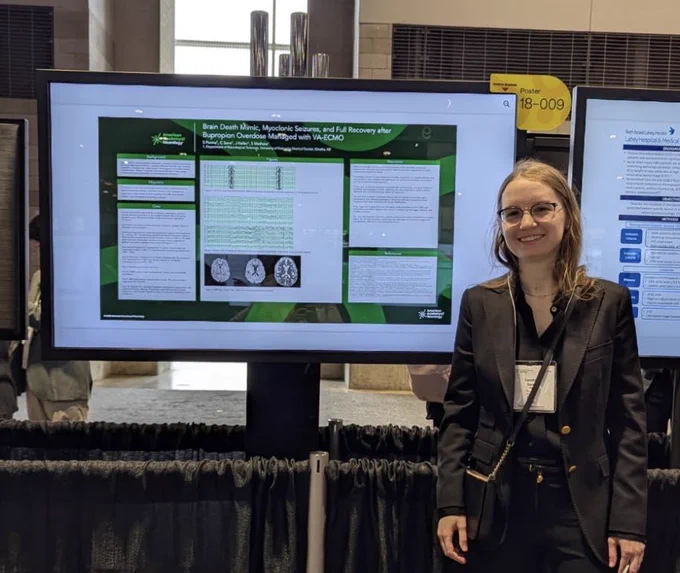 Dr. Sane dressed in business attire stands smiling beside a large digital research poster display. She is wearing glasses and a name badge. The poster behind her includes medical text, charts, and brain images, with a visible label reading “Poster 18-009.”