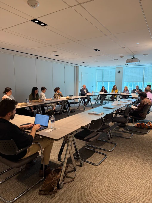 A group of people sit around U-shaped tables in a bright conference room during a discussion or panel meeting, with notebooks, water bottles, and a laptop visible in the foreground.