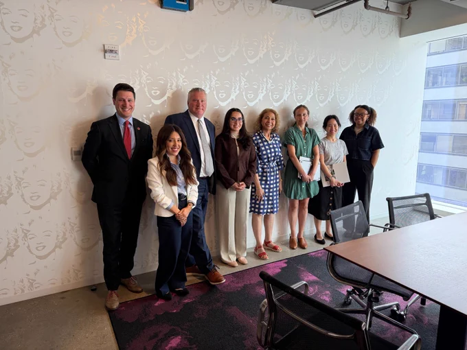 Eight people pose together in a meeting room against a patterned wall, smiling for a group photo in business and business-casual attire.