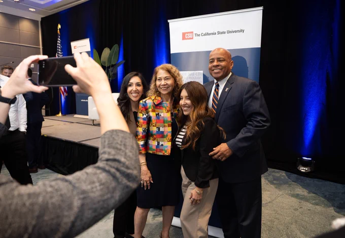 At a CSU event, four people smile and pose for a photo in front of a California State University banner while someone in the foreground takes their picture on a phone.