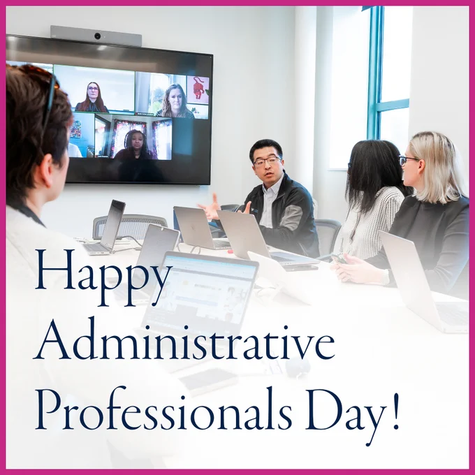 Four administrative professionals sitting together at a conference table with open laptops and a large video screen in the background showing a Zoom meeting with three more people joining the meeting remotely. With a pink border and dark navy text over a white overlay reading, "Happy Administrative Professionals Day!"