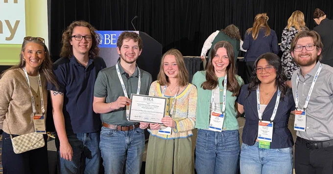 A group of seven people stand together smiling, holding a certificate. They are wearing conference badges and standing in front of a podium with a BEDA sign.