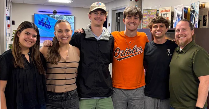 A group of six people smiling in an indoor setting. One person is wearing a shirt with the Baltimore Orioles logo. In the background, there is a screen displaying an image or video on the wall. The surroundings suggest a casual or office environment.