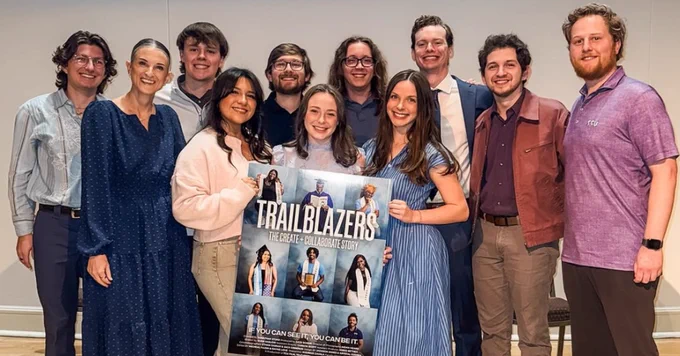 A group of people stand together indoors on a wooden floor, smiling at the camera. They are holding a "Trailblazers: The Create   Collaborate Story" poster.