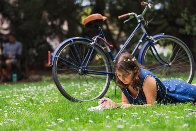 A person is lying on grass on campus, writing in a notebook. A bicycle is parked nearby. The scene is tranquil with trees and small flowers around. A blurred figure is seated in the background.