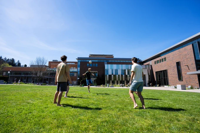 Four people are playing a game with a ball on a grassy field in front of the EMU. The sky is clear and blue.
