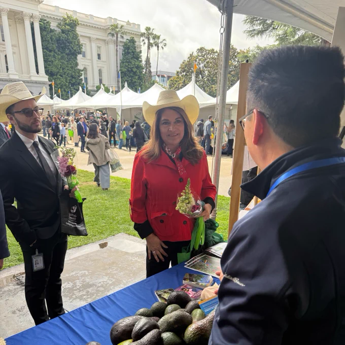 A person in a red coat and cowboy hat stands at an outdoor booth with avocados on display. Another person wearing a suit and cowboy hat holds flowers and a shopping bag. A third person, partially visible, is interacting with them from behind the booth. The setting includes white tents and a large, white building in the background, suggesting a festive or market atmosphere.