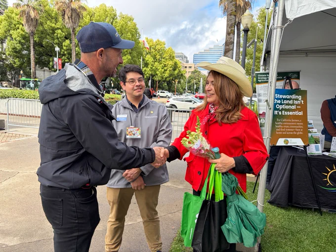 A person in a black jacket and cap shakes hands with another in a red coat and hat, holding flowers and green tote bags. A third individual stands nearby. A booth with a sign about land stewardship is in the background, with trees and buildings visible beyond.