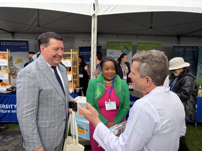 A group of individuals are gathered at an outdoor event booth featuring University of California Agriculture and Natural Resources. Banners and informational displays are visible in the background. One person is holding a white tote bag, and another is engaged in conversation while holding documents. A third person stands in the middle listening. There are also people in the background wearing hats and jackets.