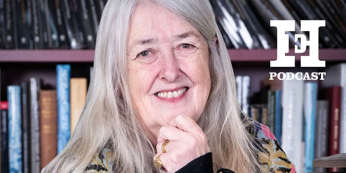 An individual with long, gray hair is smiling while resting their chin on their hand. They are in front of a bookcase filled with books. 