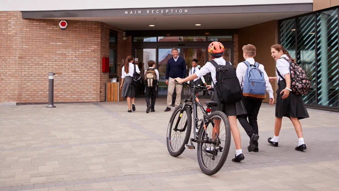 Students in school uniforms walking and biking toward the main reception entrance of a brick school building on a clear day.