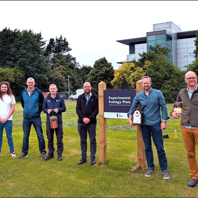 Group of six people standing near a sign labeled 'Experimental Ecology Plots' on a grassy area with trees and a modern building in the background.