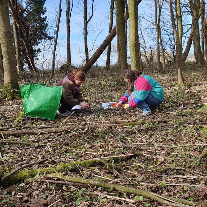 Two girls crouch in a leaf-strewn forest clearing, examining objects on the ground with a large green bag nearby.