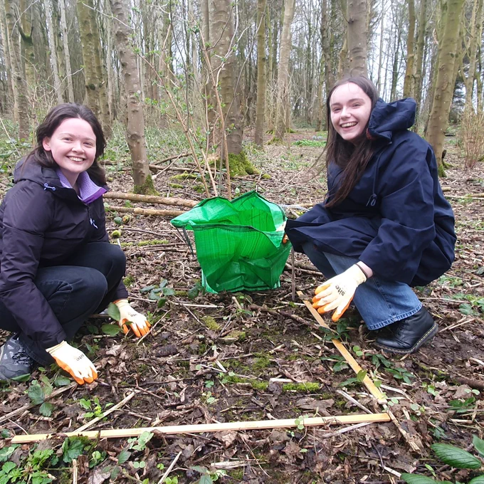 Two people wearing gloves and jackets crouch in a forest clearing, marking a square area on the ground with wooden sticks and a green bag nearby.