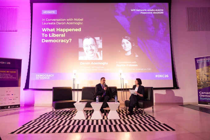 A man and a woman sitting in conversation. In the background, a projected slide reads "In conversation with nobel laureate Daron Acemoglu: What happened to liberal democracy?"