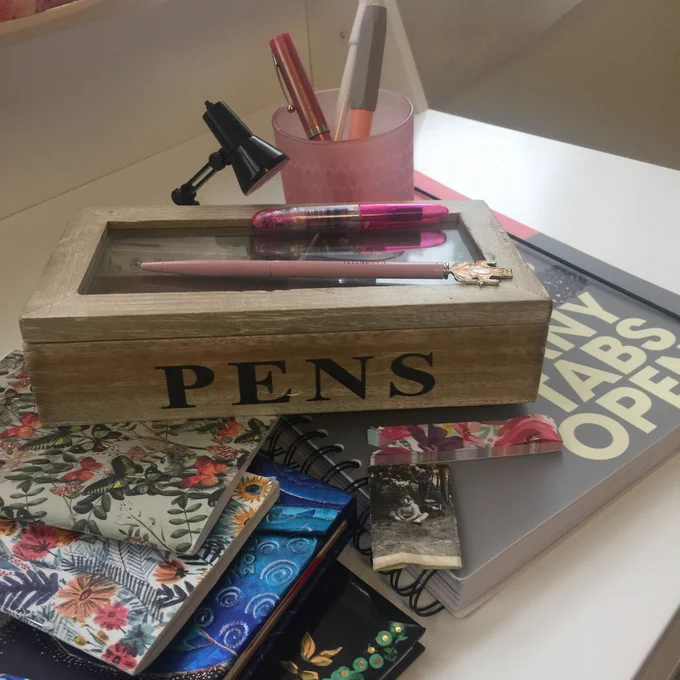An image of notebooks and pens on Penny’s desk. The notebooks are different sizes from larger A5 to slim pocket notebooks. A couple have paper covers with spindly leaves and orange flowers. One is larger with blue spirals showing and another is black with green and gold detail. A large exercise book in the right of the image reads tabs open, grey background with white text. On top of the notebooks is a wooden box with a glass lid. On the side it says pens in bold black capitals. On top are two pink pins, behind is a pink glass with two pink fountain pens and beside it a mini black anglepose desk lamp. To one side of the wooden box in the front there is a tiny black-and-white photo of Penny aged around for years old, smiling, sitting on the ground under a tree. The image is intriguing, colourful and suggestive of Penny’s work as a writer.
