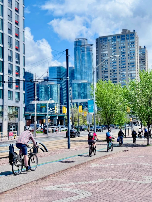 Group of cyclists riding along a city bike path with modern skyscrapers and green trees under a partly cloudy sky.