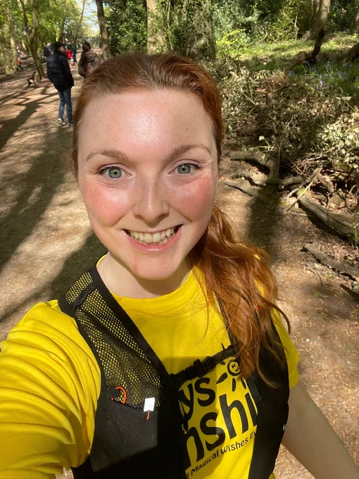 Smiling red-haired woman in a yellow charity T-shirt and running vest, taking a sunny selfie on a tree-lined woodland path.