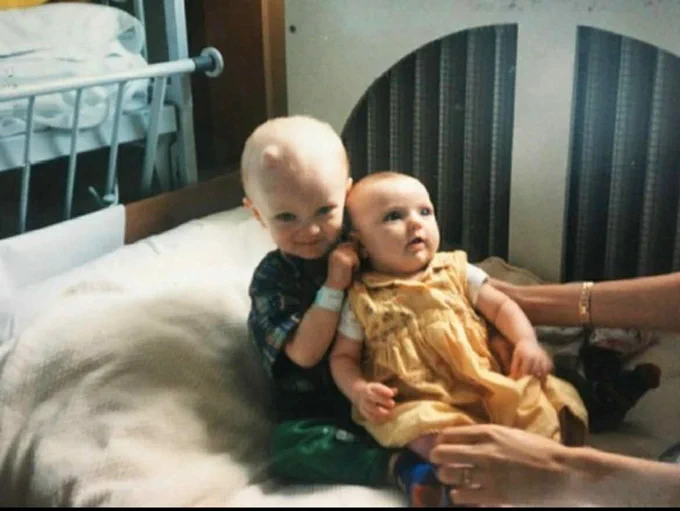 Toddler hugging an infant on a bed, adult hands steadying them; crib rail visible in the background.