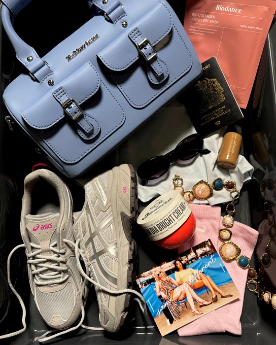 An image of a pair of beige Vagabond Hollie Sneakerinas sitting in a black tray surrounded by a taupe schuh Jaden Round Shoulder Bag, plane ticket, digital camera and accessories.