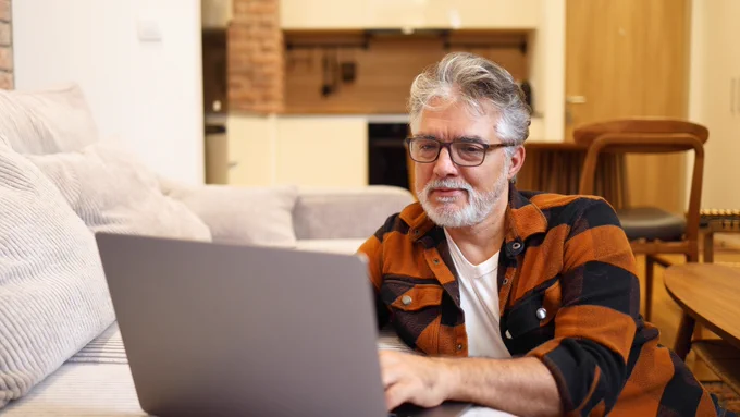 Older man with white hair wearing glasses, sitting on the floor with his laptop in front of him resting on the sofa, looking at the screen.
