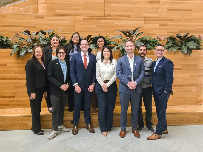 UC Merced alumni and staff dressed in professional attire posing indoors against a wooden all with green plants above.