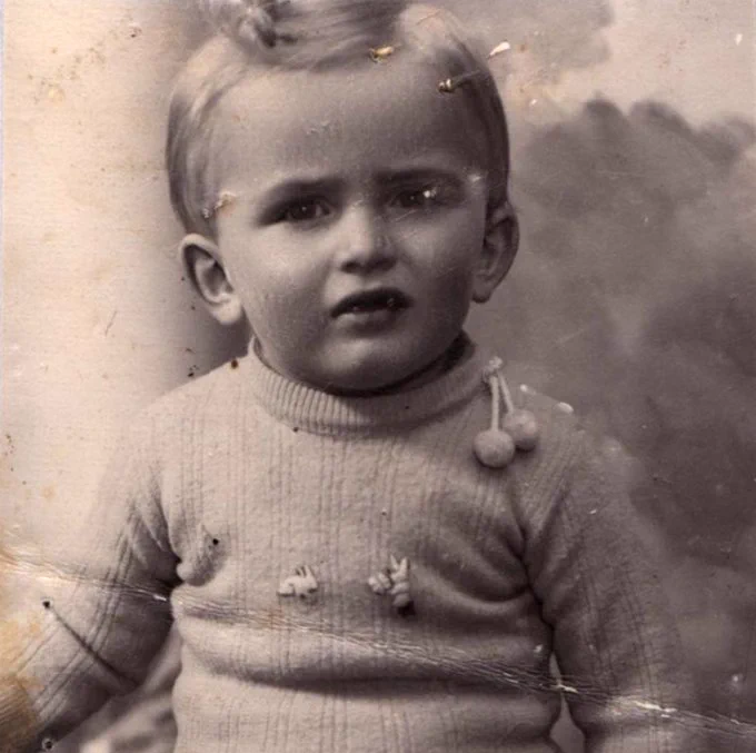 A photo of a very young boy from the waist up. He has short blond hair with a curl on top of his head. He is wearing a wool turtleneck with small pom-poms attached to the collar. 