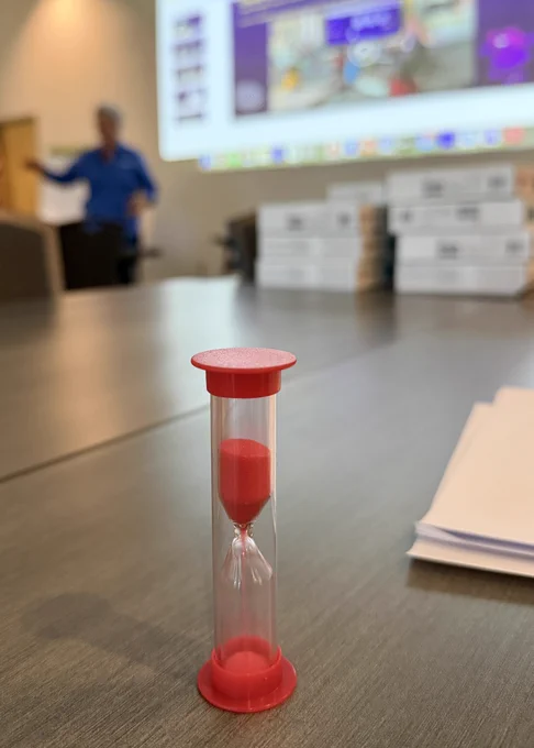 Sand timer in the main focus with a person presenting in front of a slide deck in the background
