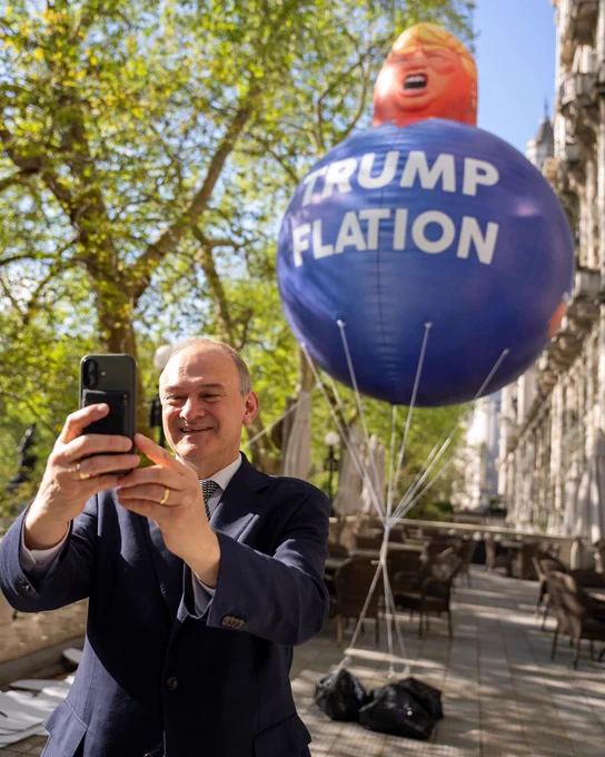 Ed Davey smiles as he takes a selfie in front of a large Donald Trump balloon reading “TRUMP FLATION”