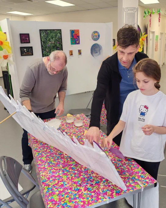 Justin and two others work together at a colourful table in an art space, carefully attaching translucent material to a long, lightweight structure, with artwork displayed on the walls behind them.
