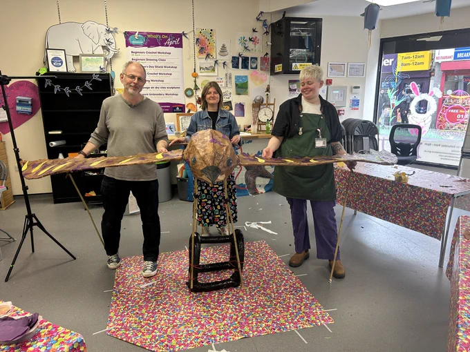 Christie, Justin and Zabby stand indoors holding a large, handmade sculptural form made from lightweight materials, with colourful artwork and creative tools visible around the room.