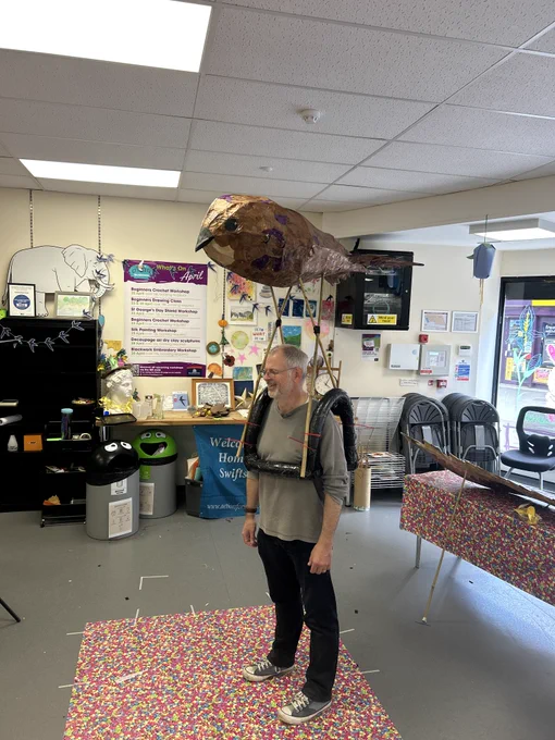 Justin stands wearing a lightweight sculptural piece suspended above their head, inside a creative workshop filled with tables, materials and artwork.