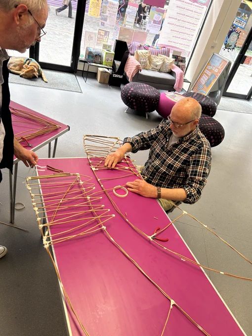Justin sits at a table carefully assembling a framework from thin wooden rods and tape, with another person standing nearby observing the process.