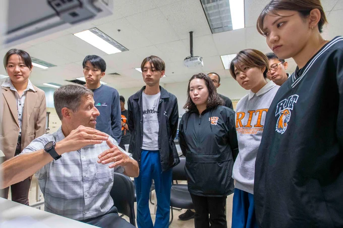 A professor sits in a chair as a table using their hands to explain something to a small group of people standing behind them