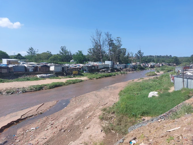 A small river running through an urban informal settlement, with makeshift homes lining the riverbank and areas of exposed soil and debris along the water’s edge.