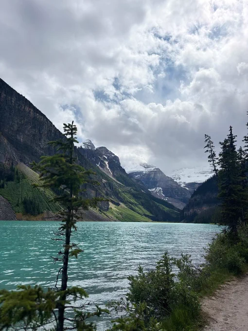 A turquoise lake surrounded by steep mountains and evergreen trees under a cloudy sky.