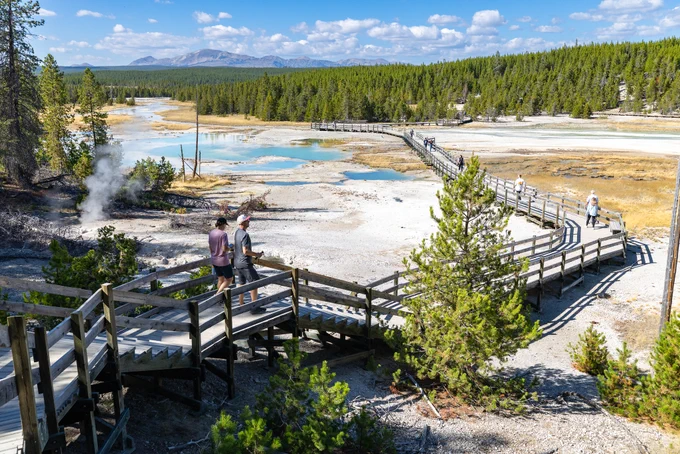 people walking on a long, narrow, wooden boardwalk that traverses safely atop a sprawling thermal feature with various colorful hot springs.