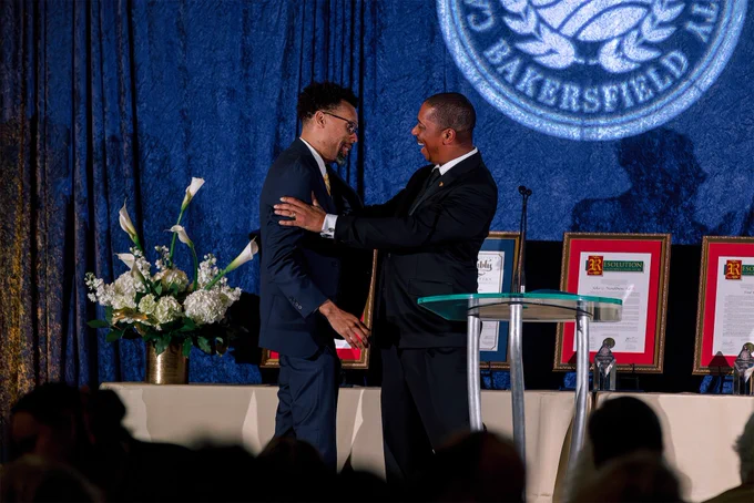 Traco Matthews (left) and President Harper (right) stand near a podium, shaking hands and smiling at each other on a stage. Behind them are framed awards and a vase with white flowers. The background features a large logo with the word "Bakersfield.