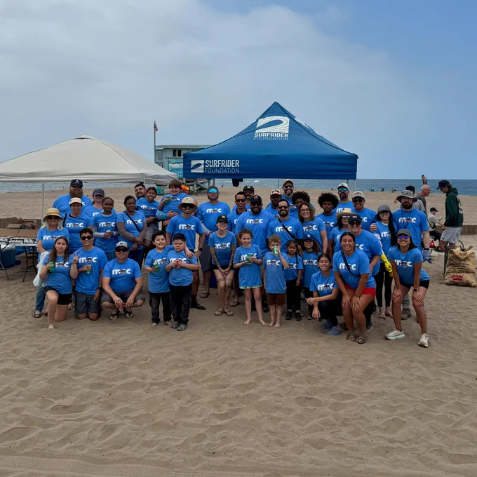 A group of people wearing matching T-shirts are gathered on a beach. Behind them are two tents and the ocean is visible in the background. There are adults and children in the group.