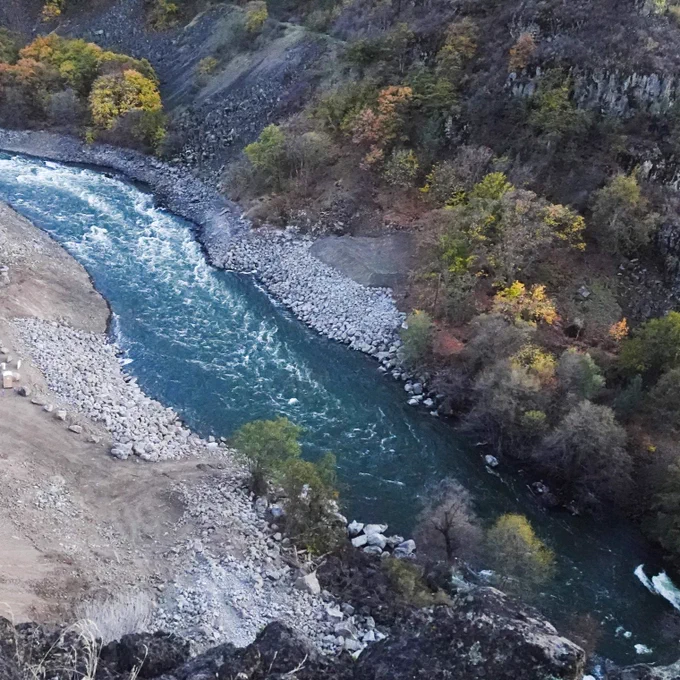 A river with clear, flowing water winds through a rocky landscape surrounded by steep, forested hills. Trees display autumn colors, creating a picturesque contrast with the dark rock formations.