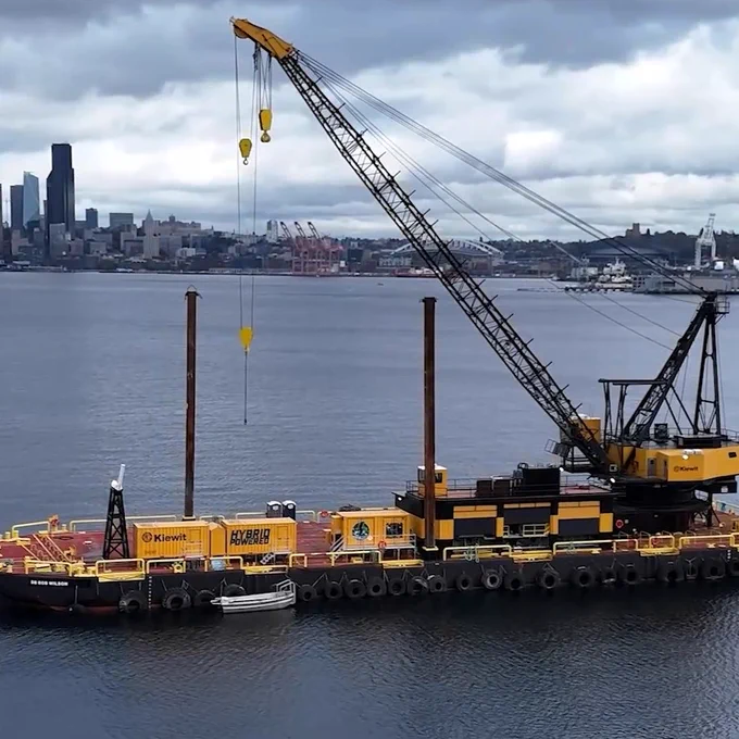 A large construction barge with a crane, labeled "Kiewit." It's floating in a body of water with a city skyline and cloudy sky in the background.