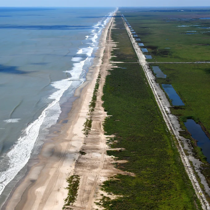 Aerial view of beach coastline, showcasing a long, narrow strip of sandy beach on the left with waves rolling in and lush green wetlands on the right. A road runs parallel between the beach and wetlands.