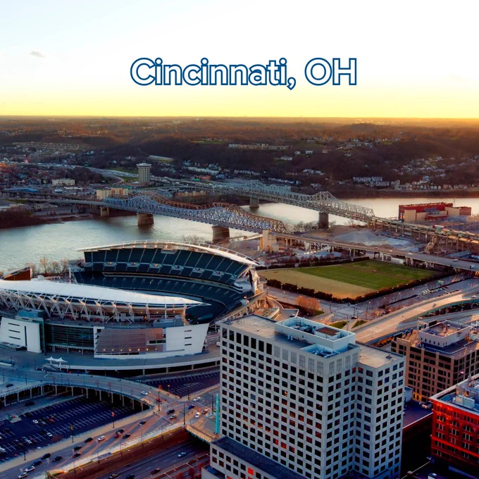 An aerial photograph of the Cincinnati, Ohio skyline during sunset. The image features the Paycor Stadium in the foreground, with several bridges spanning the Ohio River in the background. The text "Cincinnati, OH" is centered at the top in a white font with a blue outline.